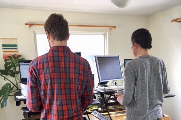 A man and a woman stand in from of a desk with their computers. The desk faces a window and their backs are to the camera.