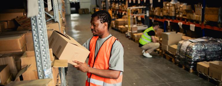 A warehouse worker in an orange safety vest handles a package in a warehouse full of packages.