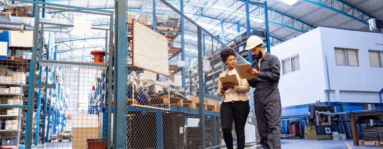 Two warehouse employees hold clipboards as they walk through a warehouse full of packages.