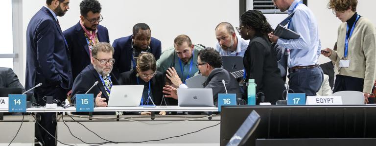 A group of people in formal apparel huddle around laptops in a white-walled room while having a discussion.