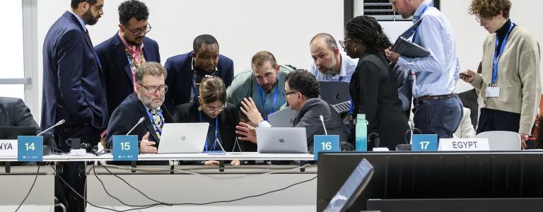 a group of people in suits huddle over a laptop in a white-walled room