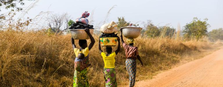 Women are walking on a dirt road in northern Benin with a bucket on their heads. A woman carries her baby on her back.