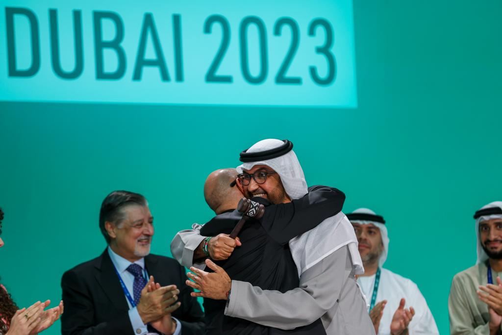 Two men, one in traditional Saudi dress and the other in a suit, embrace on the stage of COP 28 while others clap in the background