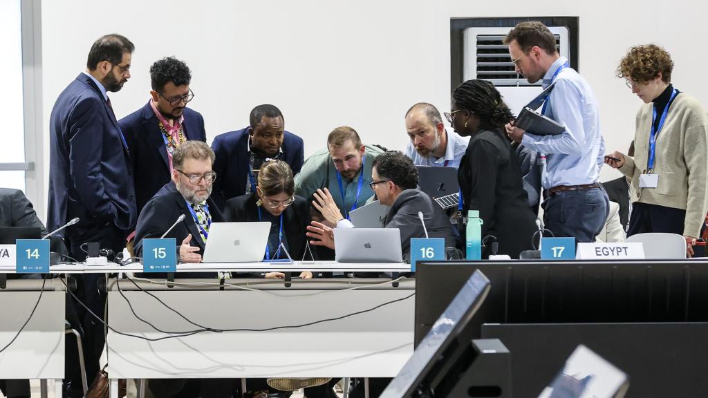 A group of people in formal apparel huddle around laptops in a white-walled room while having a discussion.